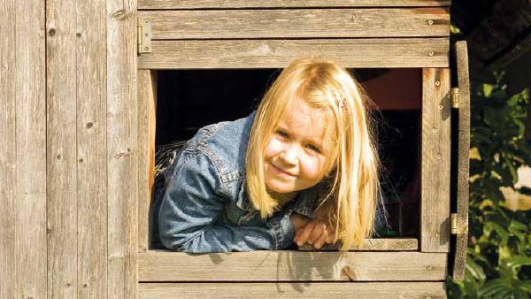Blondes Mädchen schaut lächelnd aus einem Spielhaus heraus.